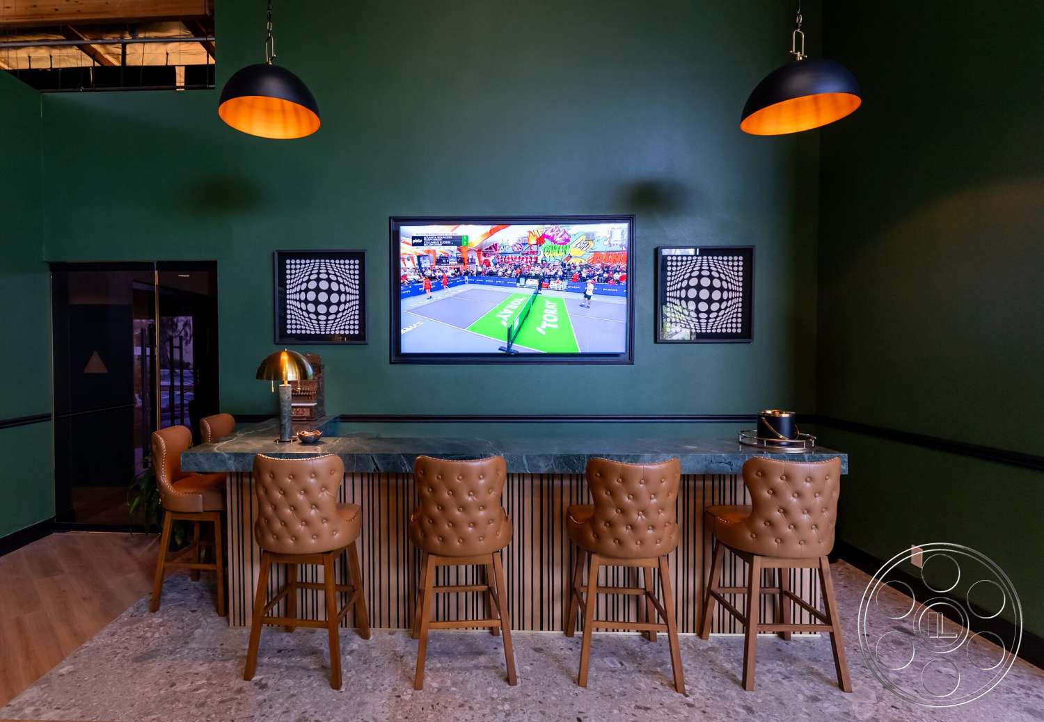 Gym 38 - light wood flooring, flat-screen television, black and white artwork, interior bar area, minimalist lighting fixtures, marble bar countertop, green painted walls, tufted leather bar stools, wooden bar front paneling