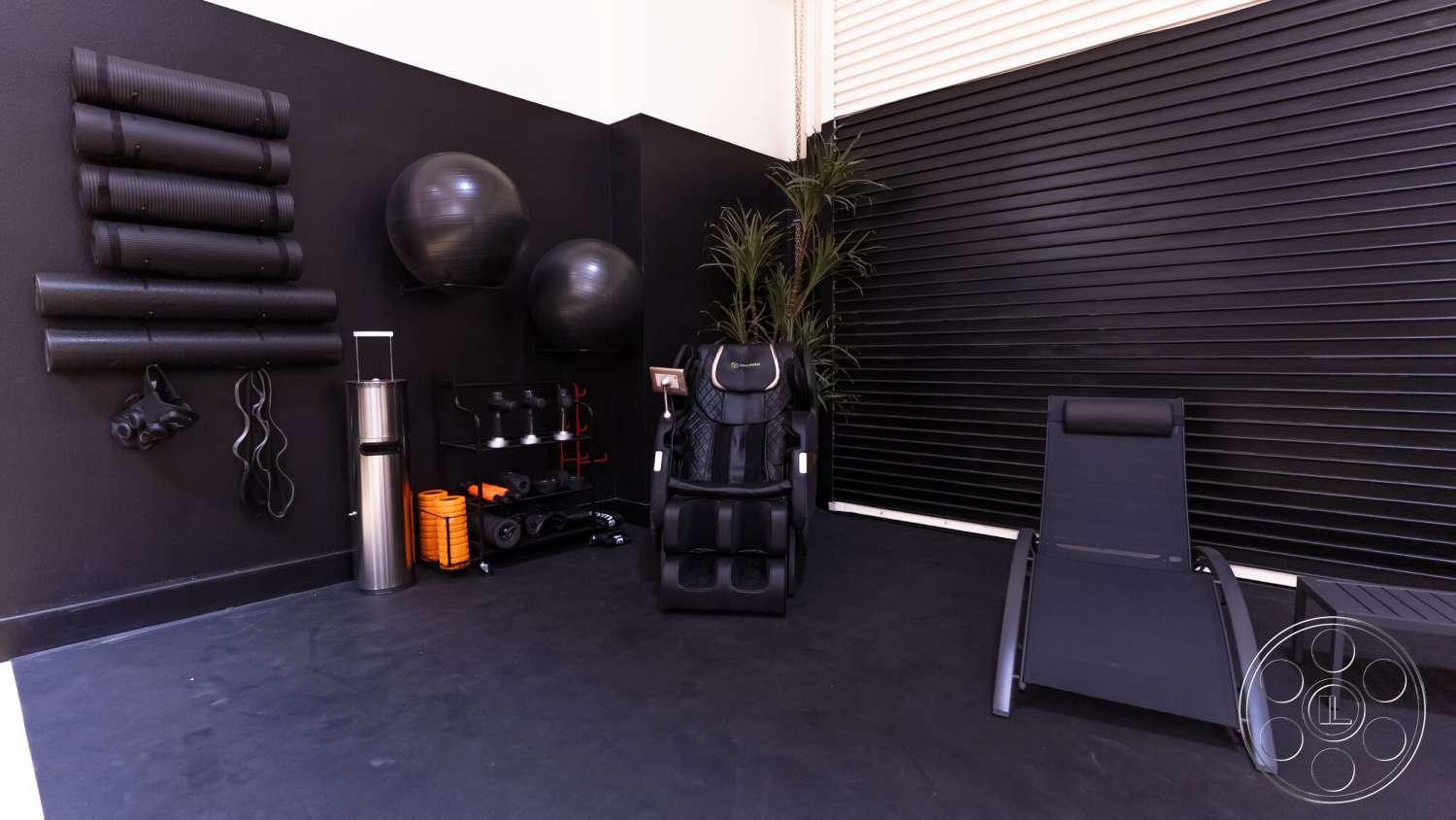 Gym 38 - modern design elements, black rubber flooring, black accent wall, interior fitness room, indoor plant decoration, exercise equipment storage rack, foldable lounge chair, black massage chair, exercise stability balls, wall-mounted yoga mats, metal water bottle holder, slatted window shades