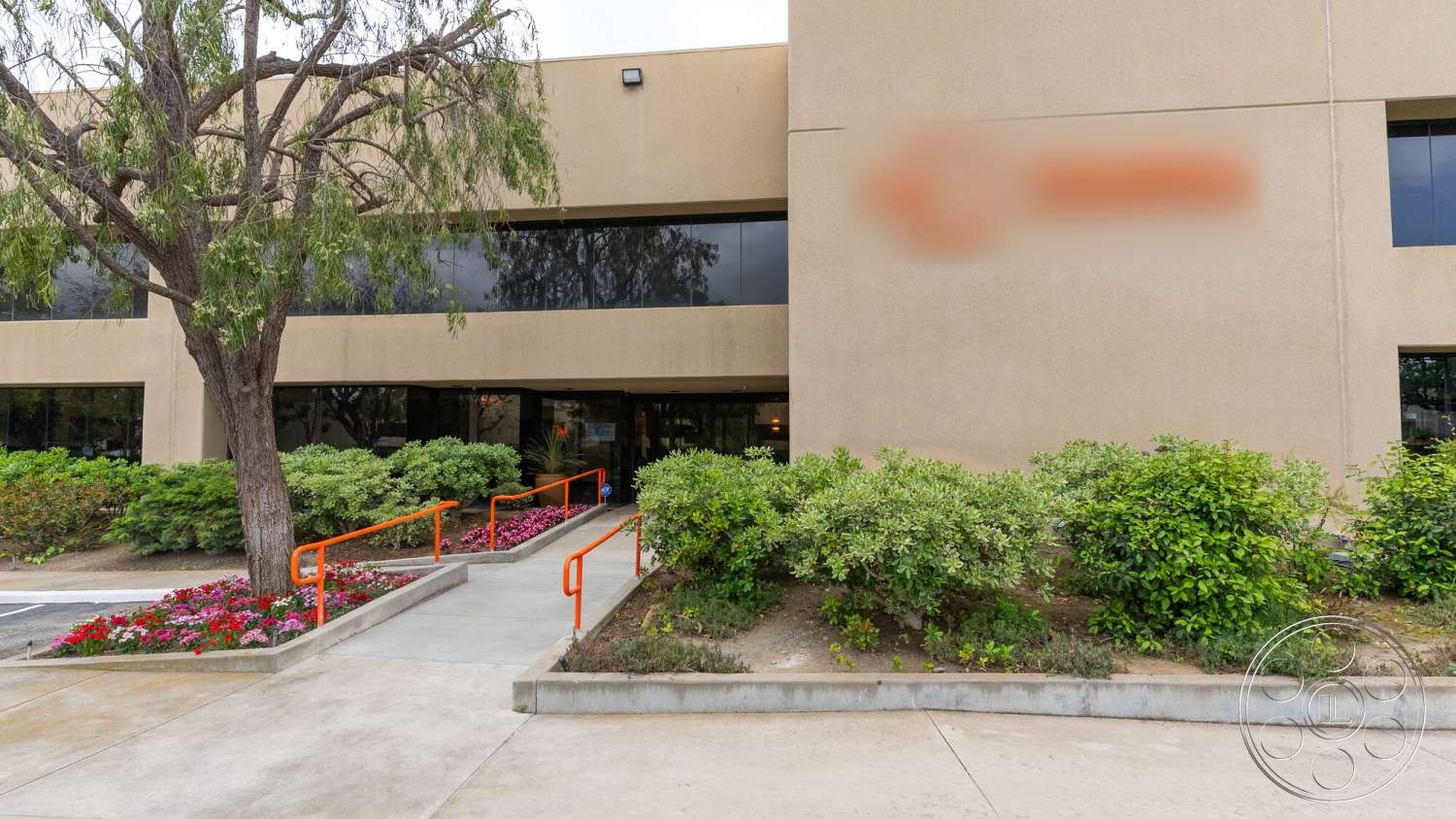 Gym 38 - exterior, landscaped garden, metal railing, large trees, light-colored stucco walls, concrete path, concrete sidewalk, brick flower bed border, glass window facades, orange metal railing, decorative flowering shrubs